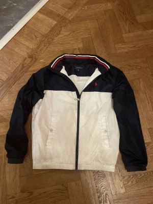 Svartvit vindjacka från Polo Ralph Lauren - Snygg vindjacka i svart och vitt från Polo Ralph Lauren. Jackan har dragkedja framtill, två fickor med knapp, och en röd logga broderad på bröstet. Kragen har en sportig randig detalj i rött och vitt. Perfekt för en cool och avslappnad stil.