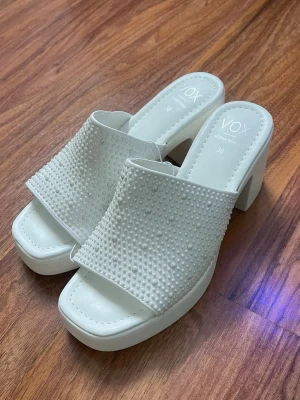 Vita sandaler med pärldetaljer VOX - Säljer ett par vita sandaler från VOX med chunky klack och öppen tå. Ovandelen är dekorerad med små pärlor för en extra snygg touch. Tillverkade i veganskt material och har en fyrkantig tåform. Perfekta för dig som vill sticka ut lite extra.