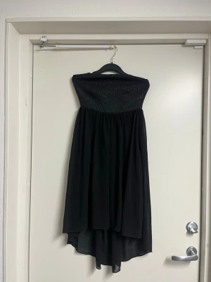 Svart bandeauklänning med asymmetrisk fåll - Säljer en svart bandeauklänning med smockad överdel och asymmetrisk fåll. Klänningen är axelbandslös och har ett flowy, lätt tyg som ger en snygg siluett. 