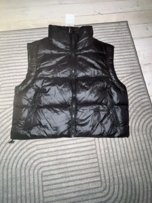 Svart dunväst från Softy, storlek S - Snygg svart dunväst från Softy med hög krage och två dragkedjeförsedda fickor. Västen har quiltad design och stängs med dragkedja framtill. Perfekt för lager på lager och ger en cool look till din outfit.