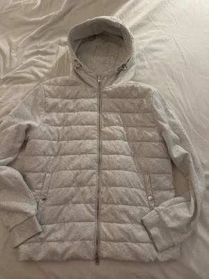 Ljusgrå quiltad hoodie med dragkedja - Snygg ljusgrå hoodie med quiltat mönster och hel dragkedja framtill. Utrustad med två fickor med tryckknappar och en justerbar huva med snören. Tillverkad i mjukt material som känns bekvämt mot huden. Perfekt för en avslappnad och trendig look.