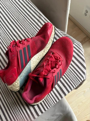 Röda Adidas Ultra Boost sneakers herr - Snygga röda Adidas Ultra Boost sneakers med svart mesh och klassiska tre svarta ränder på sidan. Ovandelen är i ventilerande mesh och sulan är vit med Boost-teknologi för extra komfort. Perfekta för dig som vill ha både stil och funktion.