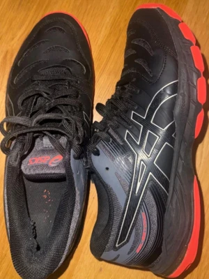 Svarta Asics sneakers med röd sula - Snygga svarta sneakers från Asics med tydliga loggor på sidorna och coola röda detaljer på sulan. Skorna har snörning och är tillverkade i en kombination av syntet och mesh för bra andningsförmåga. Perfekta för dig som gillar sportig stil.