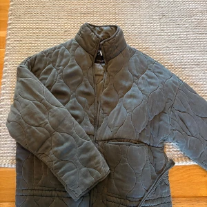 Populär Zara jacka - Snygg grå quiltad kappa från Zara manchestertyg. Kappan har hög krage, bälte i midjan och vågigt mönster i tyget. Perfekt för dig som vill ha en trendig och bekväm jacka med unik struktur.