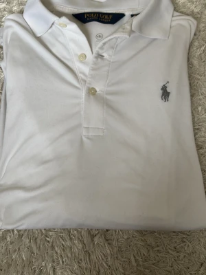 Vit långärmad piké från Polo Ralph Lauren - Klassisk  vit tunn långärmad pikétröja från Polo Ralph Lauren med knappar framtill och ikoniska loggan broderad på bröstet. Tröjan har krage och är tillverkad i mjuk bomull, perfekt för en clean och stilren look.