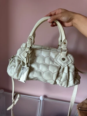 Vit quiltad handväska från Marc Jacobs - Säljer en snygg vit handväska från Marc Jacobs med quiltat hexagonmönster. Väskan har dubbla handtag, avtagbar axelrem och en metallbricka med logga. Tillverkad i läder med guldfärgade detaljer. Perfekt för dig som vill ha en unik och trendig accessoar.