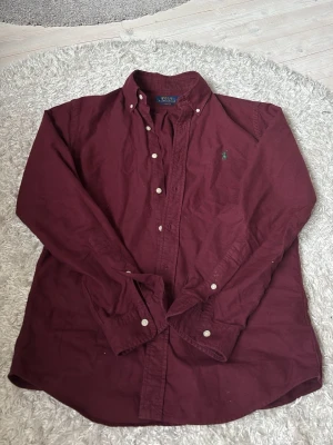 Vinröd skjorta från Polo Ralph Lauren - Klassisk vinröd skjorta från Polo Ralph Lauren med button-down krage och broderad logga på bröstet. Skjortan har vita knappar och är långärmad. Tillverkad i mjuk bomull för en bekväm känsla.