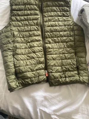 Olivgrön dunväst från Save the fuck - Snygg olivgrön dunväst från Save the duck  med horisontella sömmar och dragkedja framtill. Västen har en klassisk passform och är tillverkad i ett glansigt, quiltat material. Fjällrävens logga syns på nedre kanten bak. Perfekt för lager-på-lager-stil.