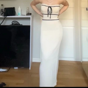 Vit långklänning med svarta detaljer - Trendig vit långklänning med svarta ränder och snörning framtill. Klänningen är axelbandslös och har en kroppsnära passform som framhäver siluetten. (Ursäkta bildkvaliteten, screenshots från en video)