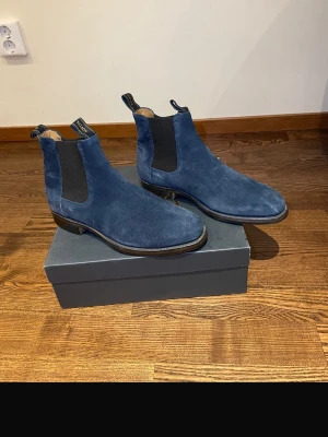 Blå mocka Chelsea boots från Loake - Snygga Chelsea boots i blå mocka från Loake. Klassisk siluett med elastiska svarta paneler på sidorna och dragflikar både fram och bak. Skorna har låg klack och svart sula, perfekta för en stilren look. Oanvända 