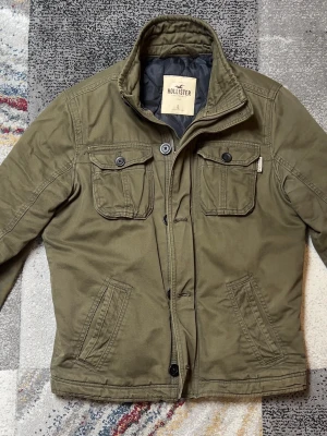 Olivgrön jacka från Hollister - Snygg olivgrön jacka från Hollister med två bröstfickor med knappar och två sidofickor. Jackan har hög krage, dragkedja och knappar framtill. Perfekt för dig som vill ha en stilren och praktisk jacka till vardags.