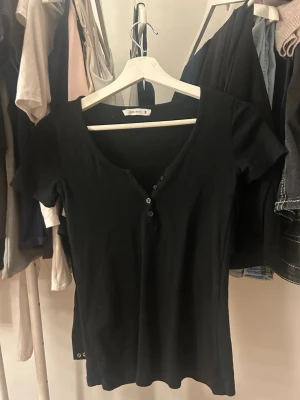 Svart topp med knappar från Terranova - En svart, kortärmad topp från Terranova i storlek S. Toppen har v-ringning med dekorativa knappar framtill och är gjord i mjuk bomull. Enkel och stilren modell som passar till det mesta.