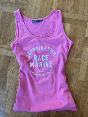 Rosa linnetopp från Race Marine - Snygg rosa linnetopp från Race Marine med vit text framtill. Toppen har rund halsringning och är ärmlös, perfekt för varma dagar. Tillverkad i mjuk bomull som känns skön mot huden. Sportig och enkel design som passar till mycket.