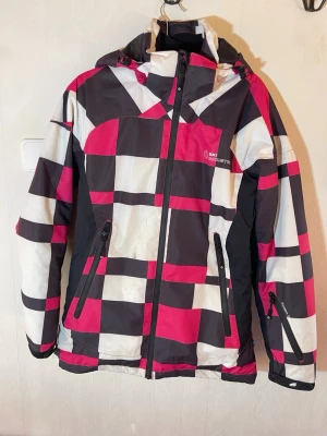 Rutig skidjacka från Ski Industries - Färgstark parkajacka från Ski Industries med rutigt mönster i grå, vit och rosa. Jackan har huva, dragkedja framtill , ficka på ärmen och två stora fickor med dragkedja. Tillverkad i slitstark syntet, perfekt för snöiga dagar. Långärmad och med sportig look. Ski Industries, storlek S. Fint skick, någon liten fäck men sparsamt använd. Djur, rök och parfymfritt hem.