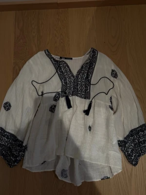 Vit boho-blus med brodyr från Zara - Säljer en luftig vit blus från Zara med mörkblå broderade detaljer och tofsar vid halsen. Blusen har v-ringning, vida långärmar och ett avslappnat fall. Materialet är tunt och skönt, perfekt för en trendig boho-look.
