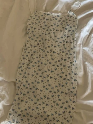 Vit blommig sommarklänning - Söt vit klänning med små blå blommor och tunna axelband. Klänningen har en rak passform och är perfekt för varma dagar. Lätt och luftig, passar till många olika stilar.