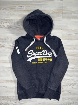 Mörkgrå Superdry hoodie med tryck - Snygg mörkgrå hoodie från Superdry med stor vit och gul logga på bröstet och japansk text🤩 Hoodien är i fint skick och passar Xs-S🙌🏻 Hör av er vid funderingar samt snabbaffär😇