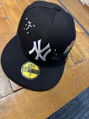 Svart New Era NY keps med brodyr - Säljer en svart New Era 59FIFTY keps med stor vit NY-logga framtill och färgglad blombrodyr på sidan. Kepsen har platt skärm och klassisk snapback-look. Materialet är bomull och polyester, perfekt för dig som vill sticka ut med snygga detaljer.
