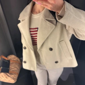 Beige skepparkavaj med knappar - Stilren beige skepparkavaj med dubbelknäppning och bred krage. Jackan har två framfickor och är tillverkad i ett mjukt ullmaterial. Perfekt för dig som vill ha en klassisk look med modern touch.