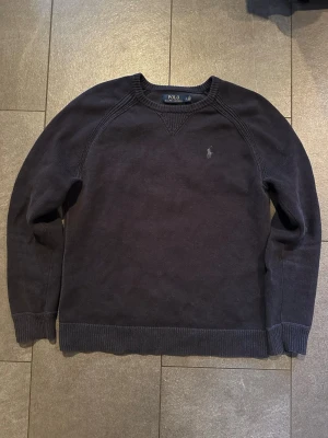 Mörkblå tröja från Polo Ralph Lauren - Mörkblå stickad tröja från Polo Ralph Lauren med rund halsringning och diskret broderad logga på bröstet. Tröjan har ribbade muddar vid ärmslut och nederkant, och är tillverkad i mjukt material som passar perfekt för lager-på-lager.