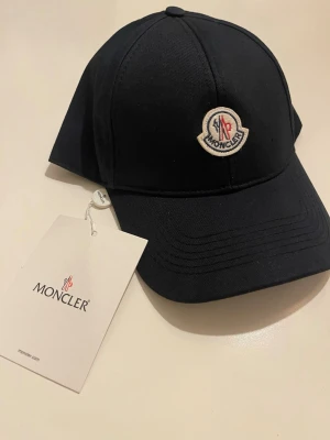 Mörkblå keps från Moncler med logotyp - Stilren mörkblå  keps från Moncler med klassisk böjd skärm och broderad Moncler-logotyp framtill. Kepsen är tillverkad i ett slitstarkt tyg och har justerbar passform baktill. Perfekt för dig som vill ha en snygg accessoar med premiumkänsla.