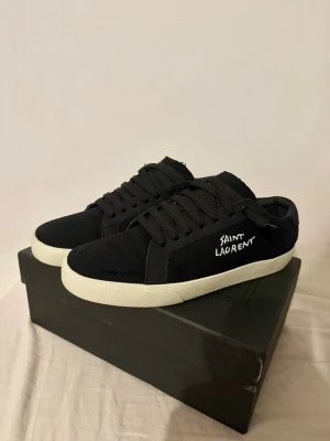 Svarta Saint Laurent sneakers - Snygga svarta sneakers från Saint Laurent med vit sula och handskriven logga på sidan. Skorna har klassisk låg profil, svarta skosnören och är tillverkade i canvasmaterial för en clean och stilren look. Ni vet varför dem är billiga