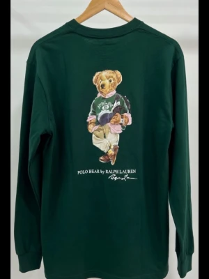 Mörkgrön långärmad Polo Bear tröja barnstorlek XL - Säljer en mörkgrön långärmad tröja från Ralph Lauren med ikoniskt Polo Bear-tryck på bröstet. Tröjan är i mjuk bomull och har en klassisk rund hals. Perfekt för dig som gillar streetwear och vill sticka ut med en cool print.