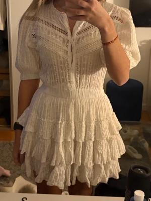 Vit volangklänning med spetsdetaljer - Supersöt vit kortklänning med volanger och spetsdetaljer. Klänningen har halvlånga puffärmar, knappar framtill och markerad midja. Materialet är lätt och luftigt, perfekt för varma dagar. Passar perfekt till studenten