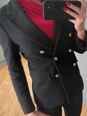 Svart dubbelknäppt kavaj med guldknappar - Stilren svart kavaj med dubbelknäppning och sex dekorativa guldknappar framtill. Kavajen har markerade axlar, breda slag och två fickor framtill. Materialet är strukturerat och ger en exklusiv känsla. Perfekt för dig som vill ha en klassisk men trendig look.