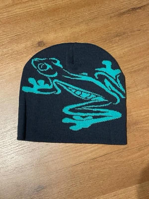 Svart mössa med turkos groda - Säljer en svart, stretchig mössa med en stor turkos groda som motiv på framsidan. Mössan är tunn och smidig, perfekt för dig som vill ha en cool accessoar med unik design. Passar både till streetwear och chill vardagsstil.