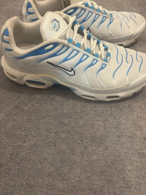 Nike Air Max Plus vita/blå sneakers - Nike Air Max Plus sneakers i vitt meshmaterial med blå detaljer och svarta swoosh-loggor på sidorna. Skorna har en sportig design med vågformade linjer och synlig Air Max-dämpning i sulan. Perfekta för dig som gillar en unik och fräsch look. Först till kvarn