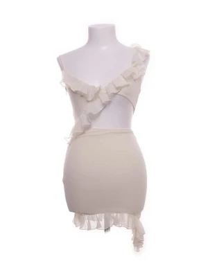 Klänning  - Trendig vit kortklänning med asymmetrisk design och volangdetaljer längs axel och nederkant. Klänningen är axelbandslös på ena sidan och har en unik, modern look som sticker ut. Perfekt för dig som vill ha något extra till sommaren.