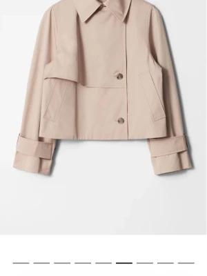 Kort beige trenchcoat  - Jätte populär Stilren kort trenchcoat från &other stories i beige med stora slag, dubbelknäppt design och breda manschetter. Jackan har två snedställda fickor och är tillverkad i ett slätt tyg som ger en klassisk look. Perfekt för dig som gillar minimalistisk och trendig stil.