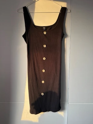 Svart ribbad klänning från SHEIN - Svart ribbad klänning från SHEIN med stora dekorativa knappar framtill. Ärmlös modell med rundad halsringning och kroppsnära passform. Perfekt för dig som gillar enkel men stilren design.