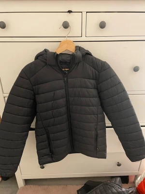 Svart pufferjacka från Everest - Säljer en svart pufferjacka från Everest med huva och dragkedja framtill. Jackan har två sidofickor med dragkedja och quiltad design som håller värmen bra. Perfekt för kalla vinterdagar och riktigt skön att bära.