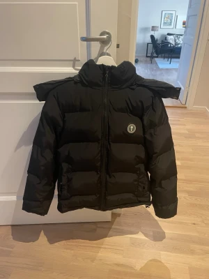 Svart pufferjacka från Trapstar - Säljer en svart pufferjacka från Trapstar med stor huva och broderad logga på ryggen. Jackan har en rund logotyp på bröstet, dragkedja framtill och breda kanaler för extra värme. Perfekt för kalla vinterdagar och riktigt snygg streetstil. 