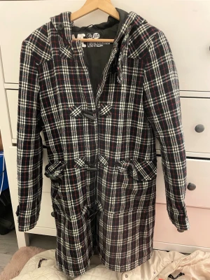 Rutig duffelcoat från Cheap Monday - Snygg rutig duffelcoat från Cheap Monday i svart, vitt och rött. Kappan har huva, klassiska träknappar med öglor och två stora fickor framtill. Perfekt för kalla dagar och ger en cool retrovibe till din outfit.