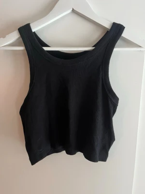 Svart ribbad croppad linnetopp - Snygg svart croppad linnetopp med ribbad struktur. Perfekt till jeans eller kjol. Ärmlös modell med breda axelband och rund halsringning. Enkel och stilren design som passar till många olika looks.