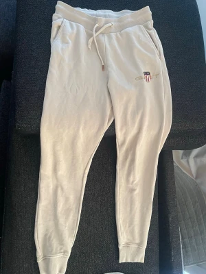 Beige mjukisbyxor från GANT - Säljer ett par beige mjukisbyxor från GANT med broderad logga. Byxorna har dragsko i midjan, muddar vid bensluten och är gjorda i mjukt bomullsmaterial. 