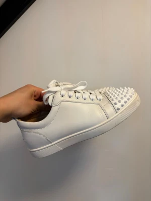 Christian Louboutin jr sneakers vita - Säljer ett par vita sneakers från Christian Louboutin i läder med coola vita nitar på tån. Skorna har klassisk låg profil, vita skosnören och en stilren design som sticker ut. Perfekta för dig som vill ha något extra till din outfit.