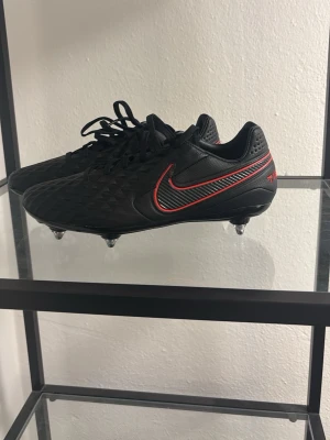Nike Tiempo fotbollsskor  - Säljer ett par HELT NYA svarta Nike Tiempo fotbollsskor med röda detaljer. Skorna har snörning,läder och fasta skruv dobbar. Tveka inte med å höra ev er vid frågor! Påsen ingår självklart!