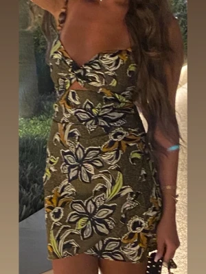 Blommig kort klänning med cut-out från Zara  - Säljer en snygg kort klänning med blommigt mönster i grönt, gult och vitt. Klänningen har smala axelband, omlott-detalj vid bysten och en liten cut-out framtill. Perfekt för sommarens alla tillfällen!