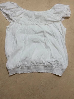 Vit offshoulder topp med spetsvolang - Söt vit offshoulder topp med bred spetsvolang runt axlarna och resår i nederkant. Toppen är ärmlös och har en luftig passform, perfekt för varma dagar. Tillverkad i mjuk bomull som känns skön mot huden.