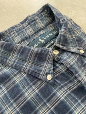 Blårutig skjorta från Ralph Lauren - Kortärmad skjorta från Ralph Lauren i blått rutigt mönster. Skjortan har klassisk krage, knäppning framtill och är tillverkad i mjuk bomull. Perfekt för en avslappnad och stilren look.