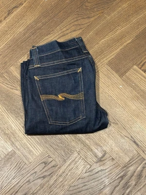 Mörkblå jeans från Nudie Jeans - Säljer ett par mörkblå jeans från Nudie Jeans med klassisk femficksmodell och kontrastsömmar i gult. Jeansen har rak passform och är tillverkade i rå denim för en stilren look. Perfekta för dig som gillar tidlös och clean stil. Storlek 30/32