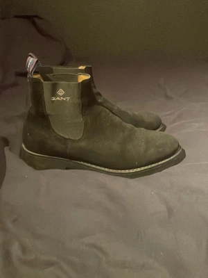 Svarta Chelsea boots från Gant - Snygga svarta Chelsea boots från Gant i mocka med klassisk siluett och elastiska paneler på sidorna. Skorna har en diskret logga på hälen och dragflikar med randigt mönster för enkel påtagning. Perfekta för dig som gillar stilrena och tidlösa skor.