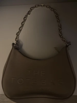 Beige The Tote Bag axelremsväska - Säljer en beige handväska från Marc Jacobs med texten 'The Tote Bag' präglad på framsidan. Väskan har en chunky silverfärgad kedjerem och är tillverkad i ett slätt konstlädermaterial. Perfekt för dig som gillar trendiga accessoarer.