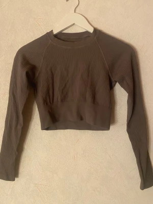 Sport ribbad crop topp - Snygg mörkgrå croppad topp med lång ärm och ribbad struktur. Tillverkad i stretchigt syntetmaterial, perfekt för träning eller chill. Rund halsringning och bred mudd nedtill. Liten logga baktill.