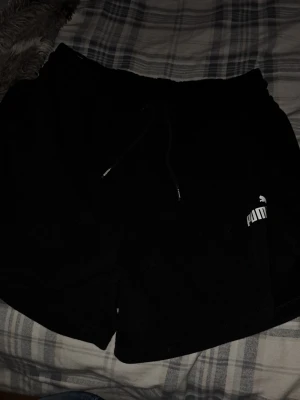 Svarta shorts från Puma med snörning - Svarta shorts från Puma med elastisk midja och snörning framtill. Klassisk Puma-logga i vitt på ena benet. Sköna och sportiga, perfekta för chill eller träning. Tillverkade i mjuk bomull för extra komfort.