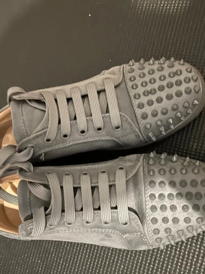 Grå sneakers med nitar - Säljer ett par grå sneakers med coola nitar på tån. Skorna har en låg profil, rund tå och är gjorda i mockaliknande material. Matchande grå skosnören och en stilren design som sticker ut med sina metalliska detaljer framtill.